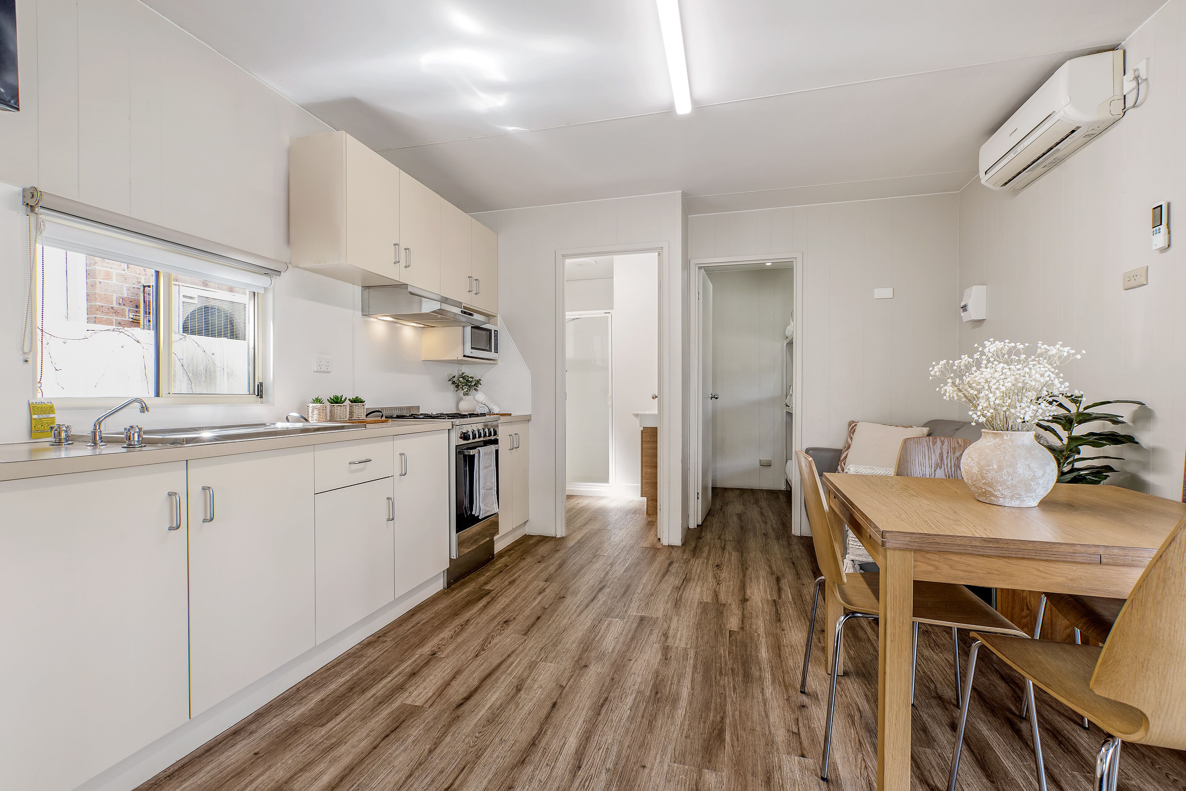 A modern kitchen with white cabinets, a wooden dining table, and a view into the living area.