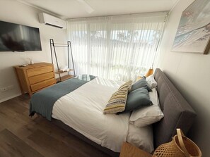 Een slaapkamer met een bed, een houten dressoir, een televisie en een raam met dunne gordijnen.