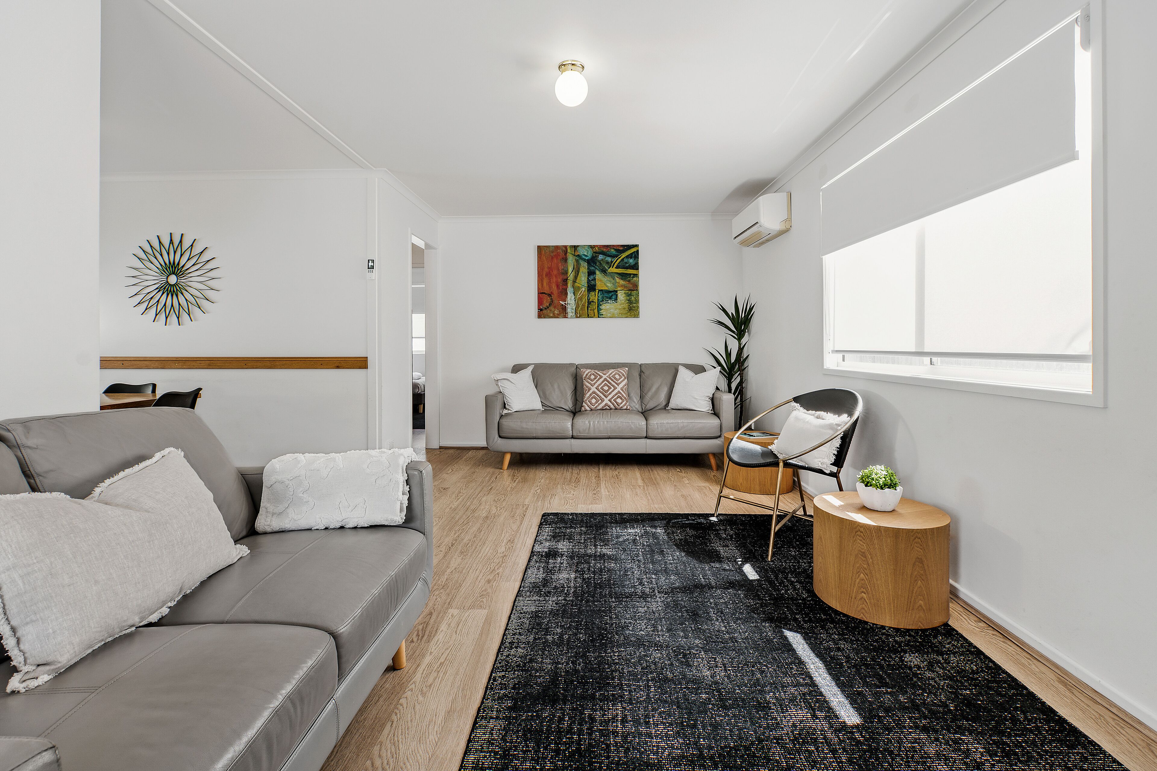 Un salón moderno con un sofá gris, un sillón gris, una alfombra negra, una mesita de madera con una planta y un cuadro abstracto colorido en la pared.