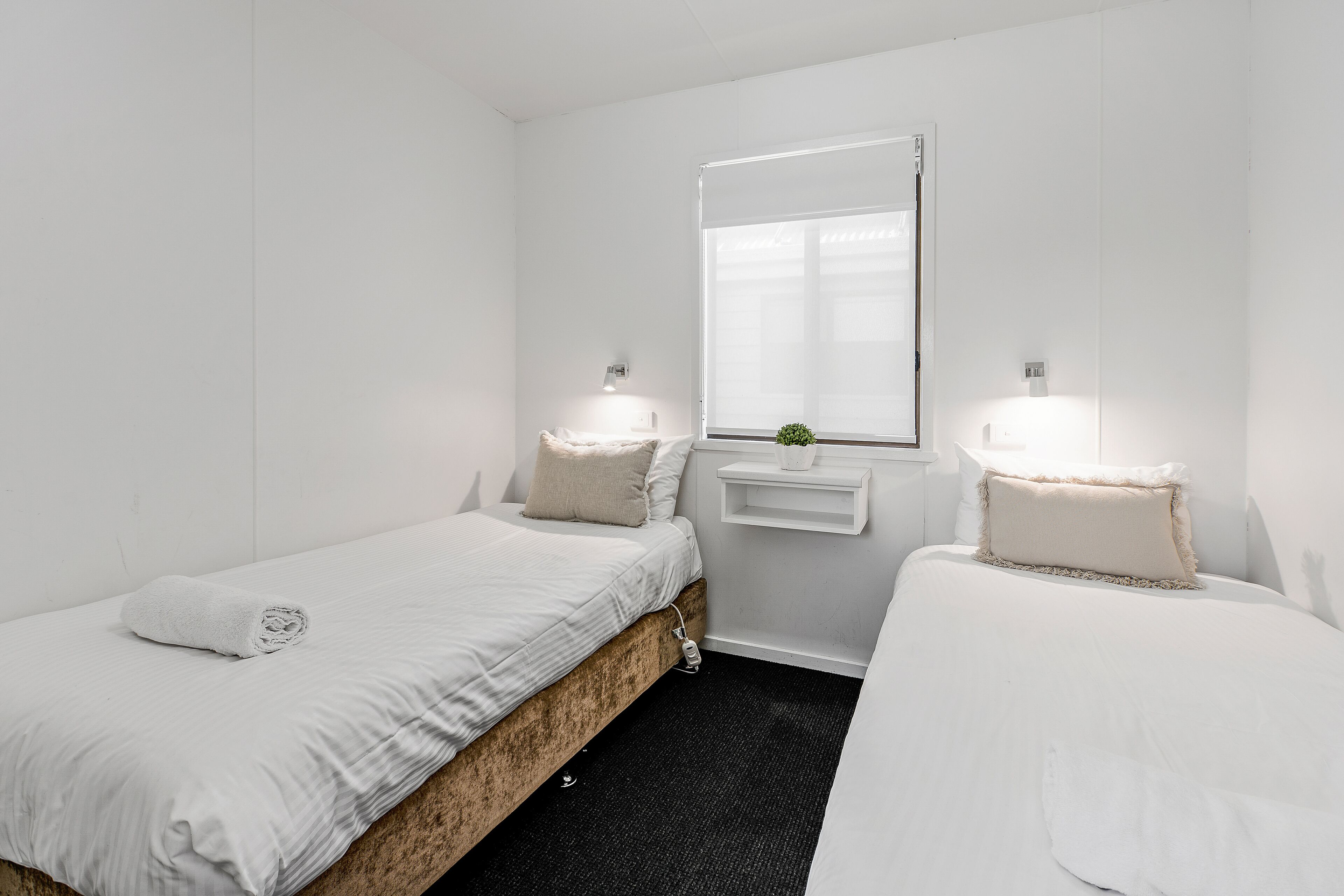 Ein kleines, sauberes Zimmer mit zwei Einzelbetten, einem Fenster mit Jalousien und einem kleinen Regal mit einer Pflanze.