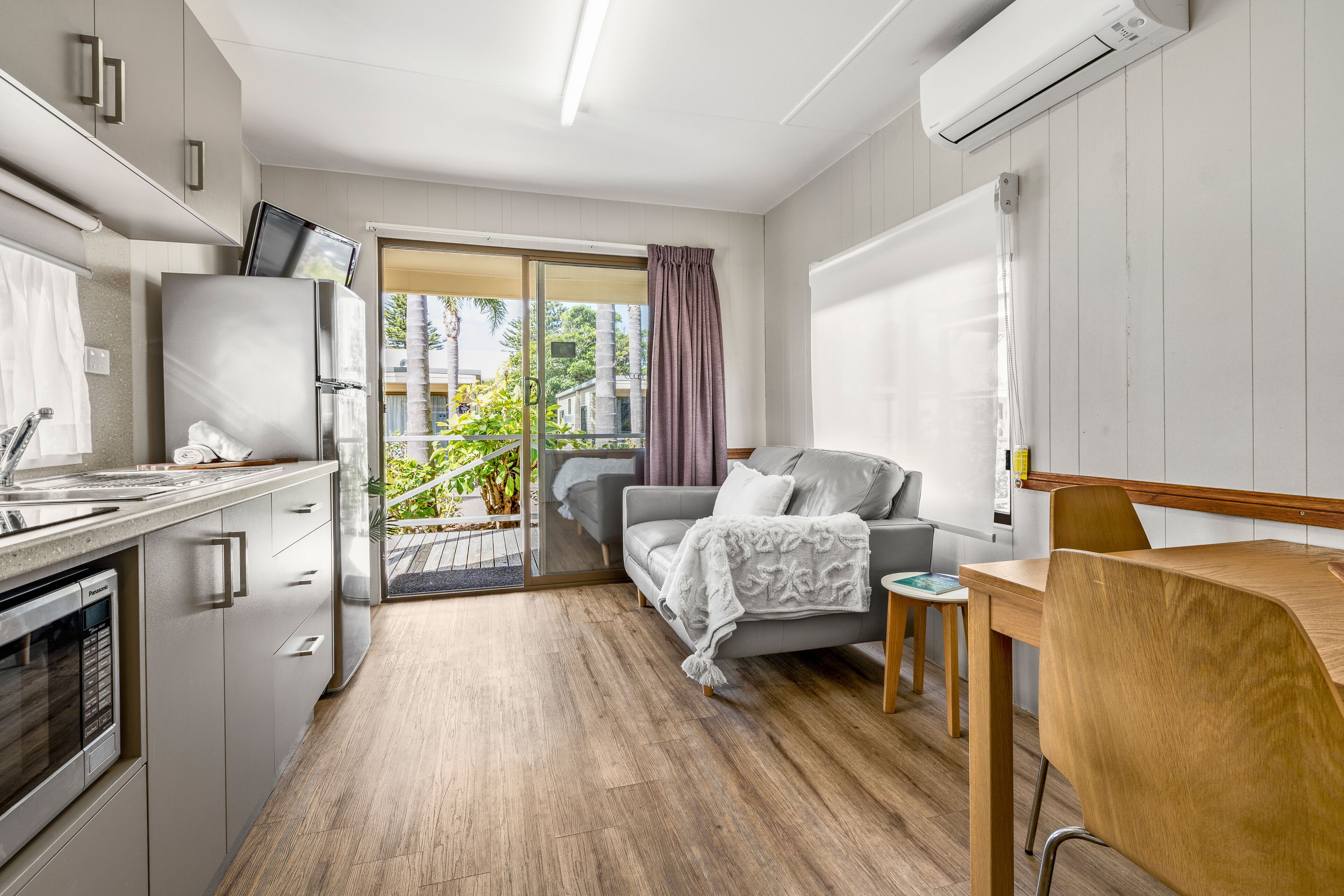 A modern kitchen with a built-in oven, a refrigerator, and a dishwasher. There is a dining area with a wooden table and chairs. The room has a sliding glass door leading to a balcony with plants and a view of buildings.