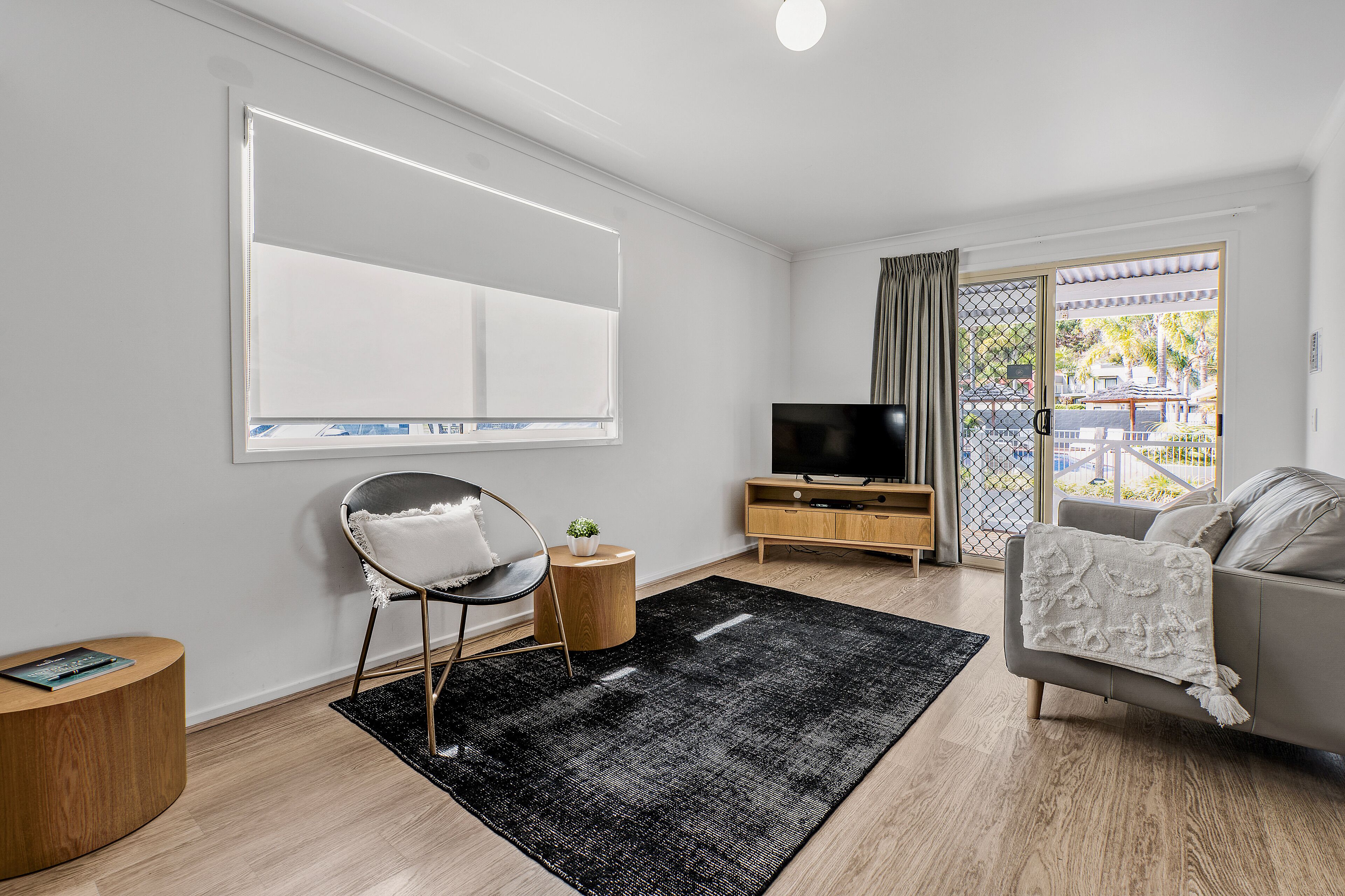 A modern living room with a flat-screen TV, a grey sofa, a round wooden side table, and a black rug.