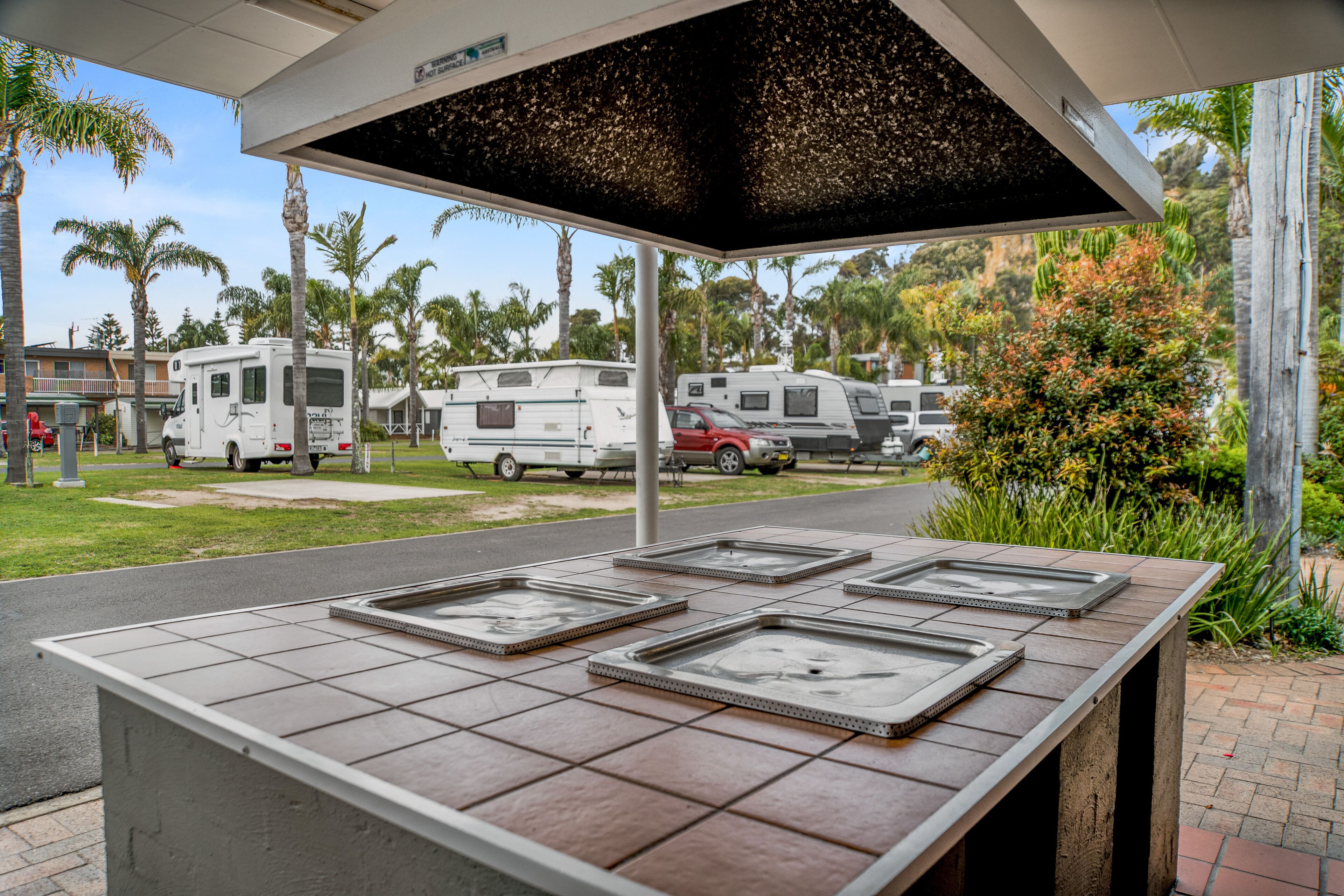Een overdekt buitengebied met een betegelde tafel waarop lege bakplaten staan, omringd door palmbomen en campers op de achtergrond.