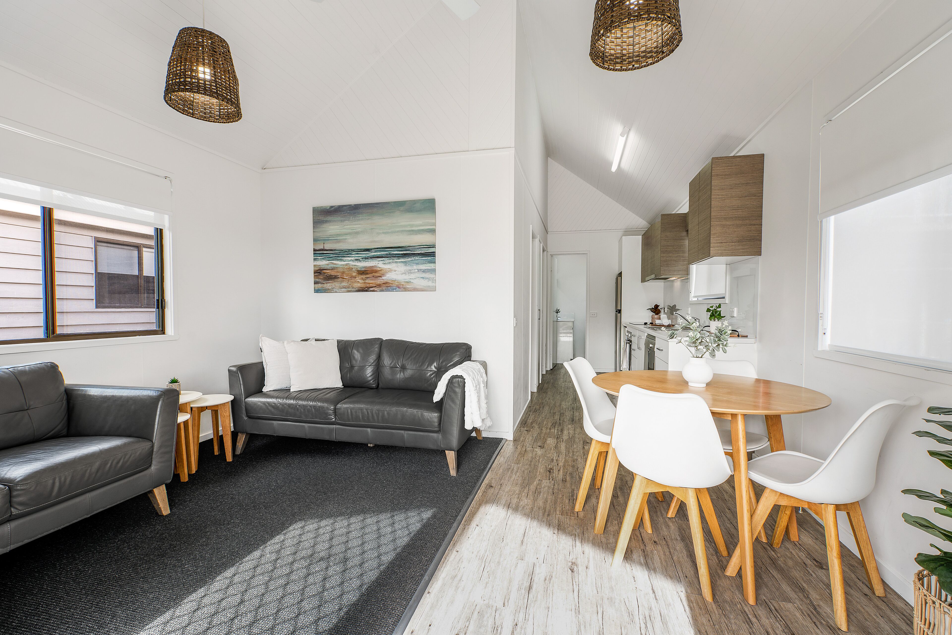 A modern living room with a grey leather sofa, a small round table, and white chairs. There is a painting on the wall and a wicker pendant light.