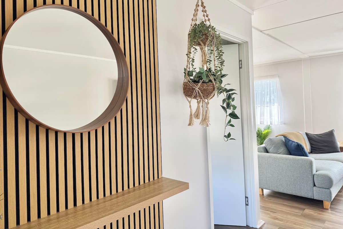 Un salon moderne avec un grand miroir rond, un mur recouvert de lambris en bois, une plante suspendue en macramé et un canapé gris.