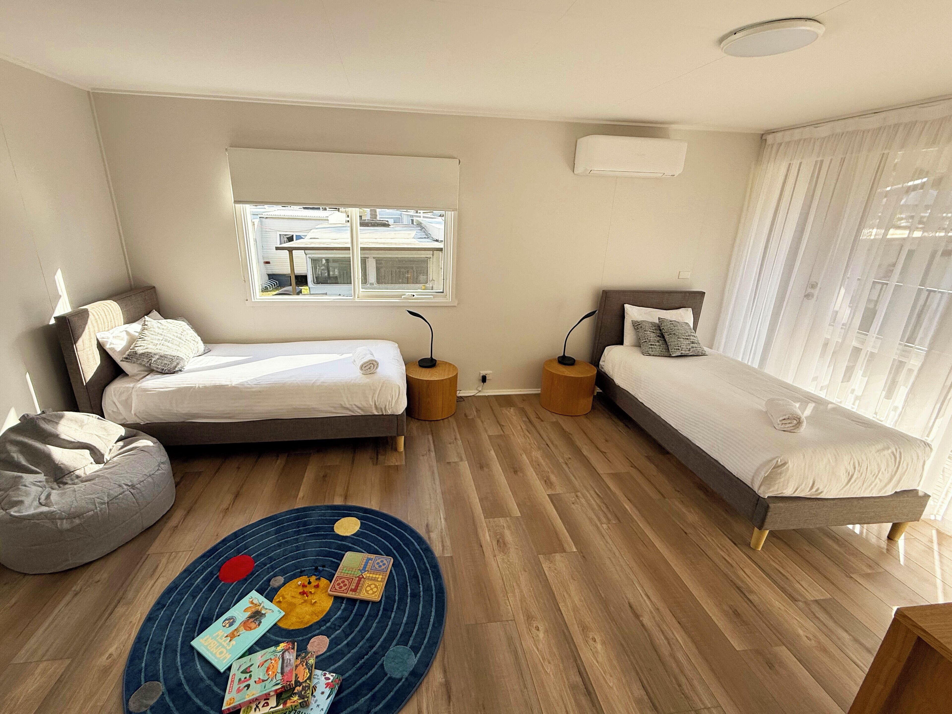 A modern bedroom with two beds, a round rug with children's toys, and a window with blinds.