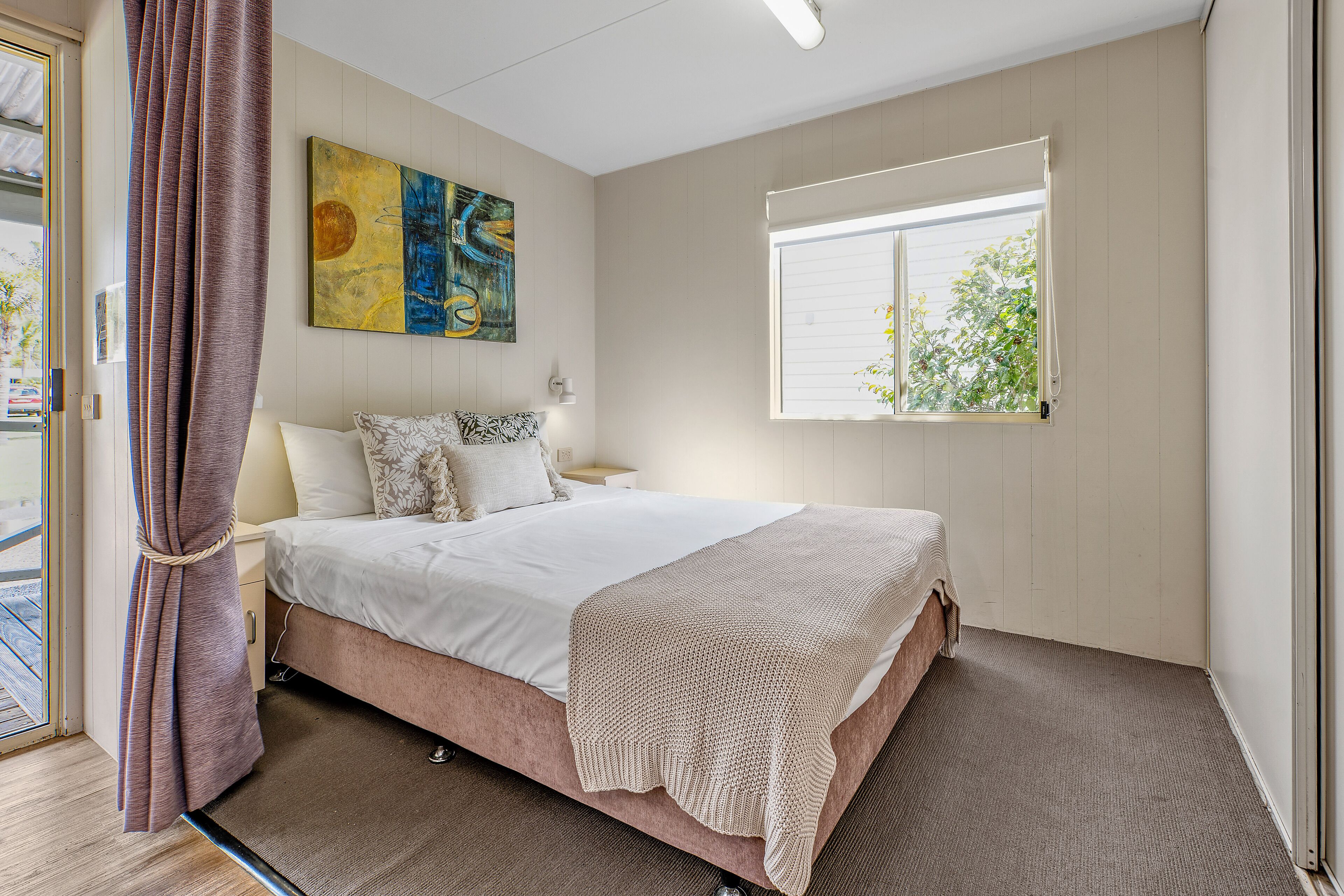 Un dormitorio con cama, ventana con persianas, un cuadro en la pared y una cortina.