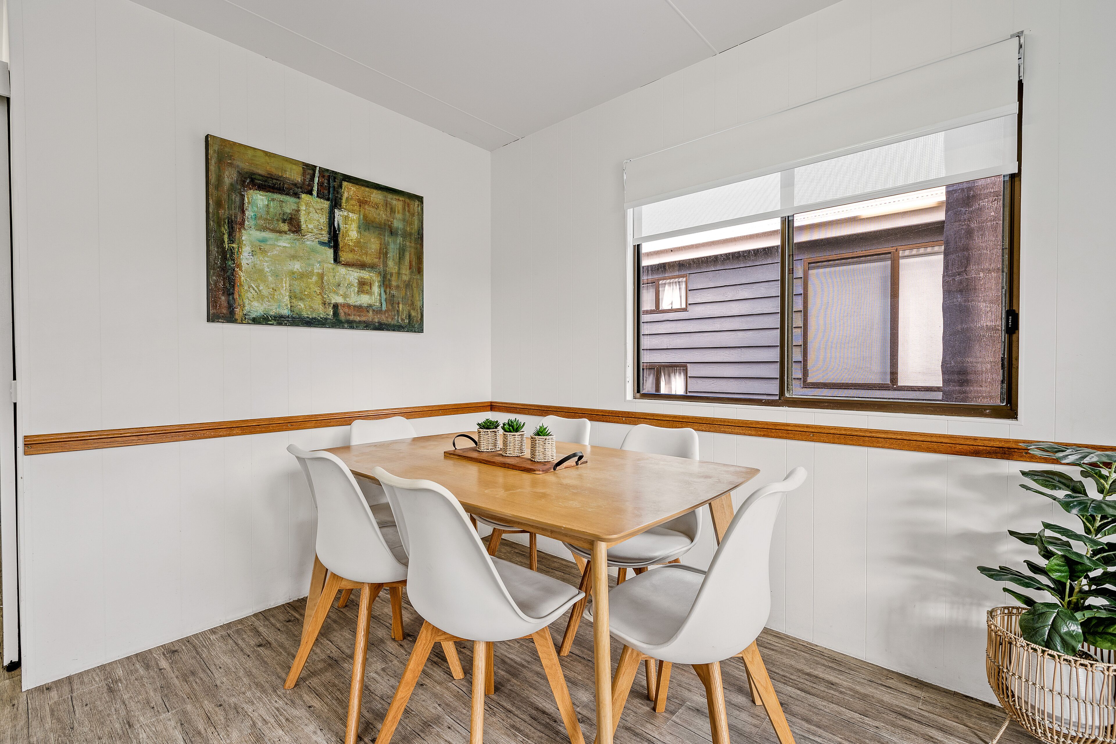 A dining area with a wooden table and white chairs, a painting on the wall, and a window with blinds.