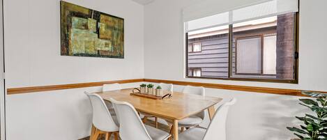A dining area with a wooden table and white chairs, a painting on the wall, and a window with blinds.