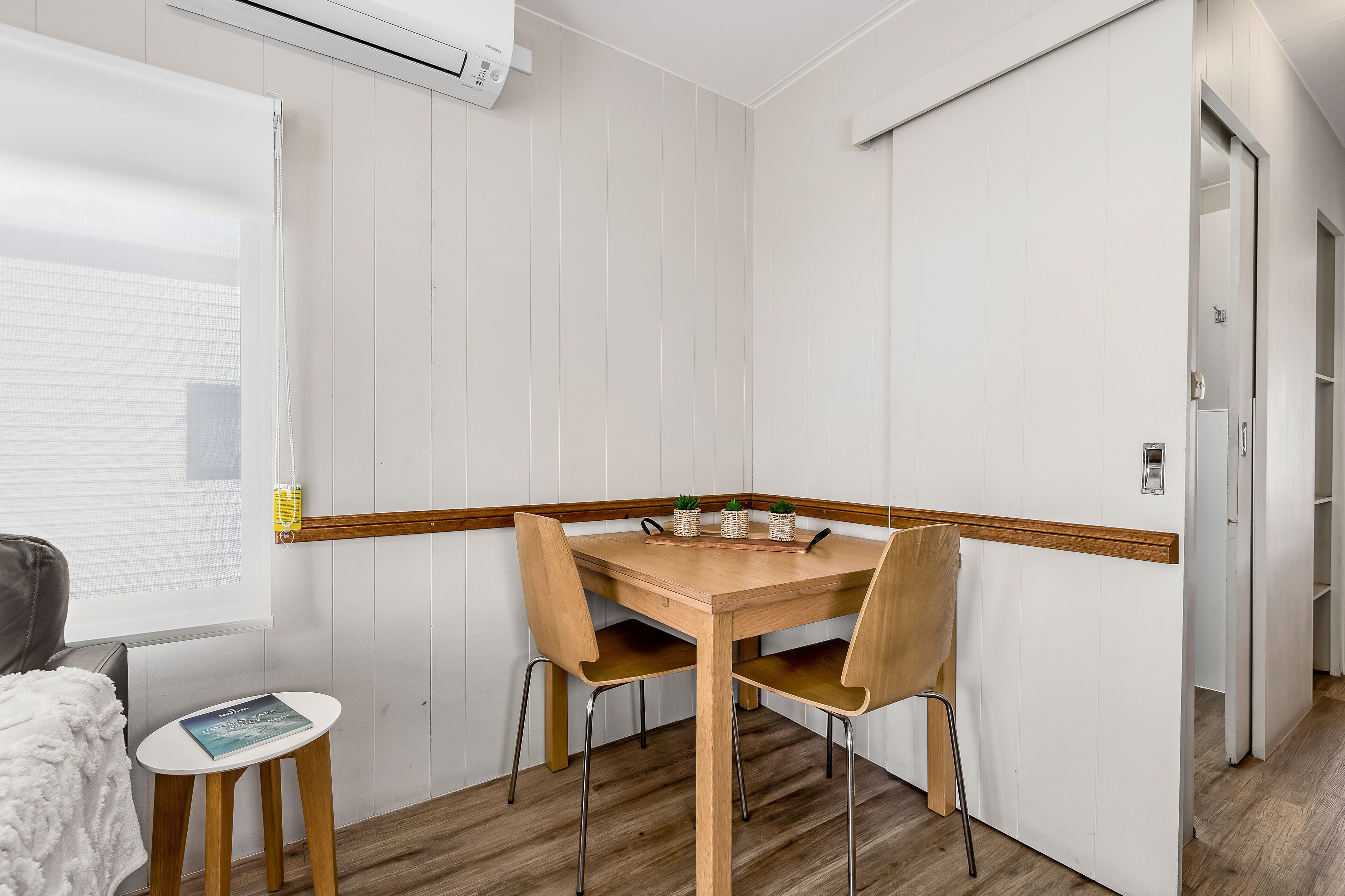 A small, neatly arranged room with a wooden table and chairs, a small side table with a book, and a window with blinds.