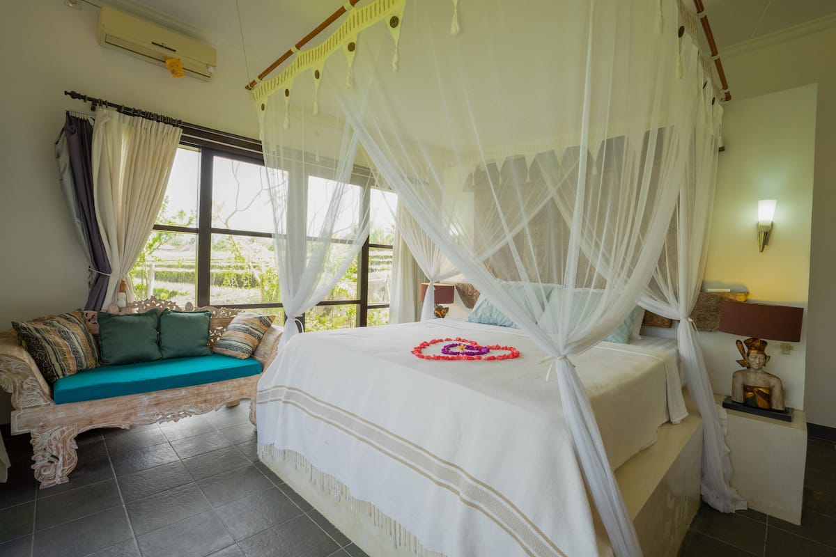 A bedroom with a bed covered by a white canopy, a sofa with cushions, a statue on a nightstand, and a window with curtains.