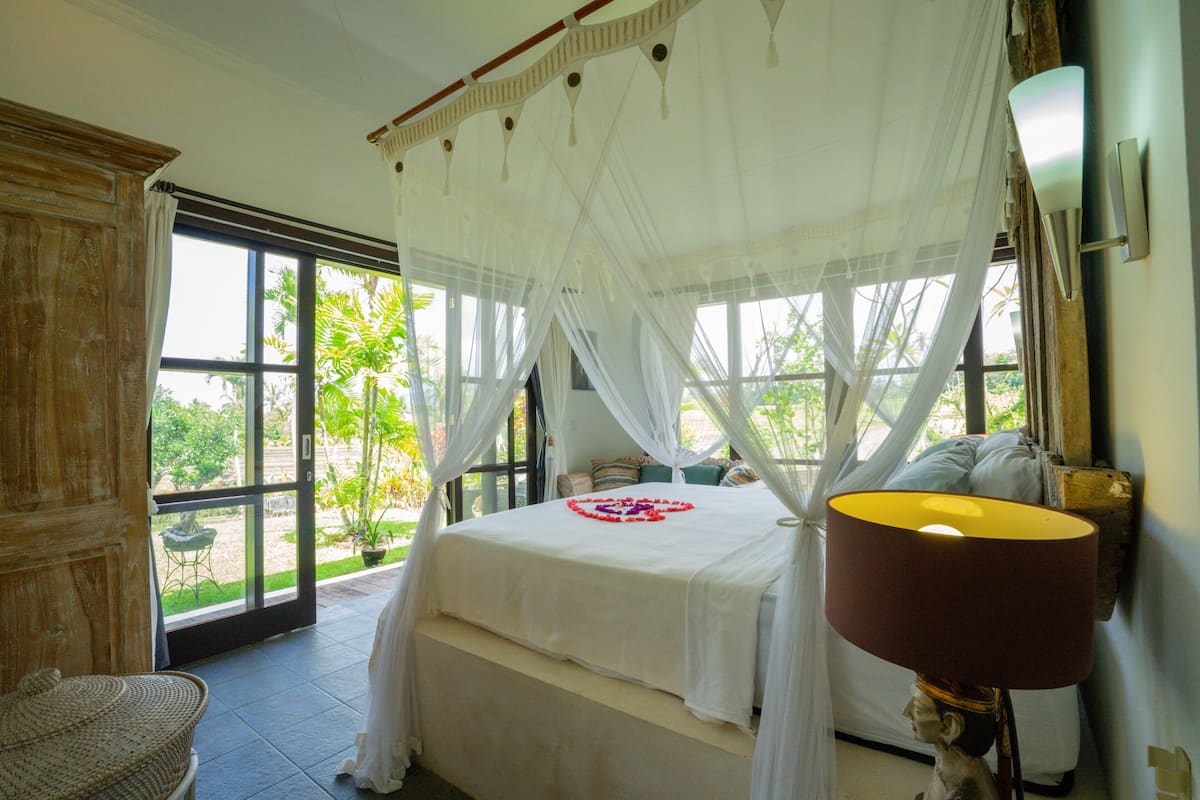 A bedroom with a four-poster bed, a large window, and a nightstand with a lamp.