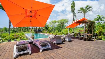 Terraza de madera con sillones de descanso y un paraguas naranja, piscina y plantas tropicales.