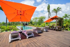 Terraza de madera con sillones de descanso y un paraguas naranja, piscina y plantas tropicales.