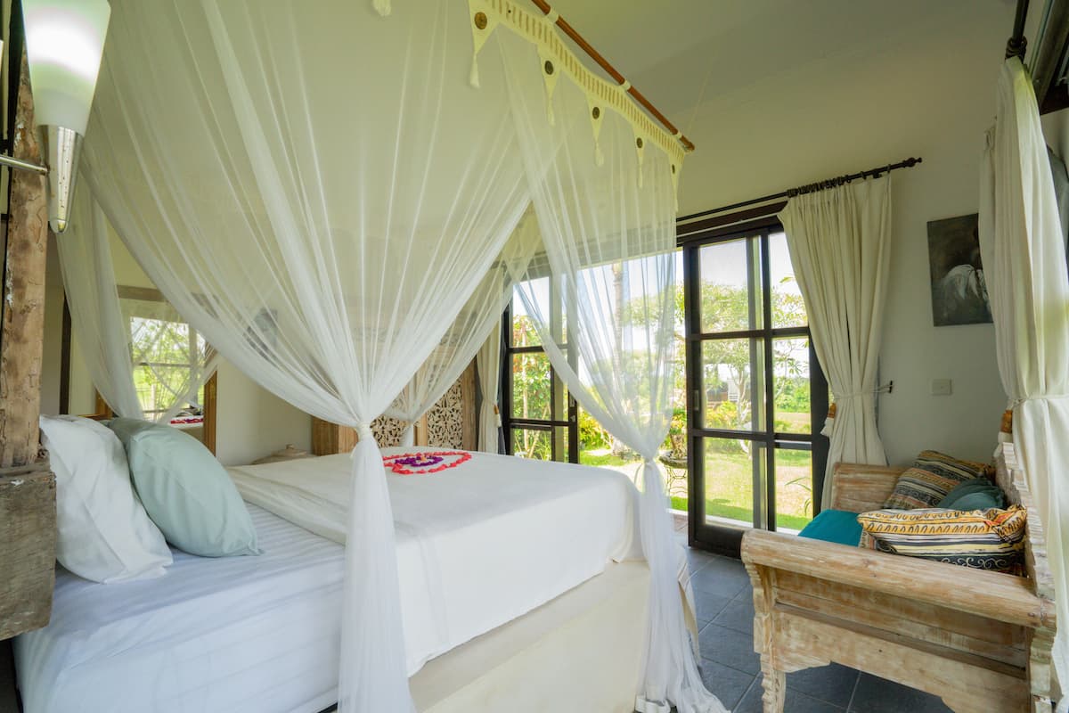 A bedroom with a four-poster bed, a wooden bench, and a view of greenery outside.