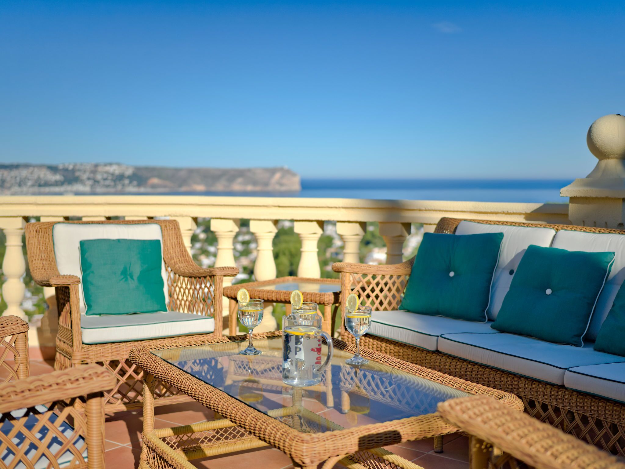 A wicker outdoor seating area with a glass table, cushions, and glasses of water.