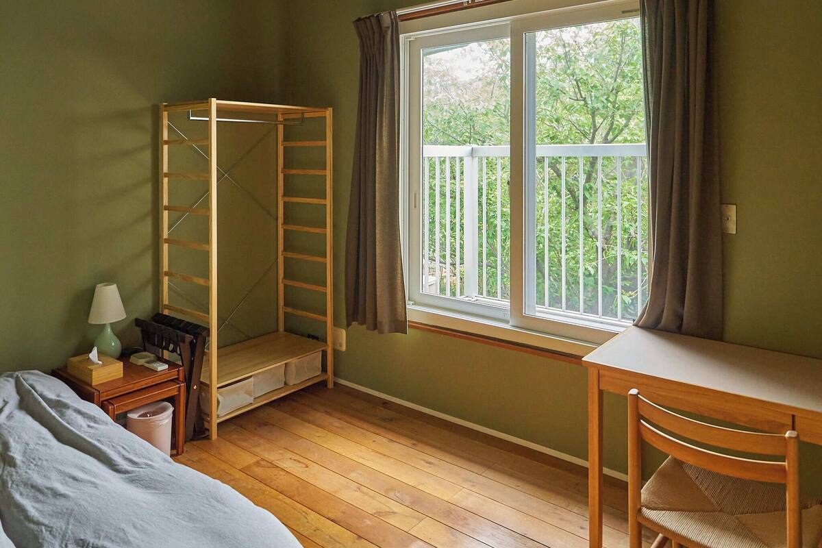 A room with a wooden bunk bed, a desk with a chair, a window with a view of greenery, and a small table with a lamp.