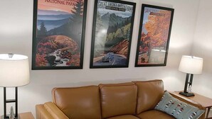 A tan leather sofa with a cushion, flanked by two wooden side tables with lamps, against a white wall with three framed posters of Great Smoky Mountains National Park. - Sleeps up to 8 in a safe, quiet area (Kodak)