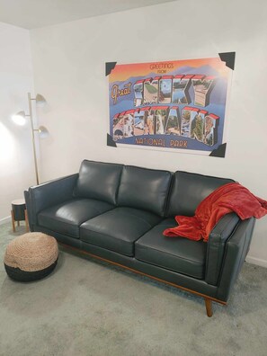 Een zwarte leren bank met een rode plaid, een rond geweven poef, en een poster op de muur die Great Smoky Mountains Nationaal Park promoot.