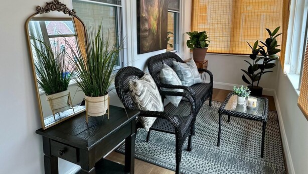 A cozy living room with a dark wooden coffee table, a wicker sofa with patterned cushions, a large mirror, and potted plants. - Boston Hospitality Homes, Harvard (Boston)