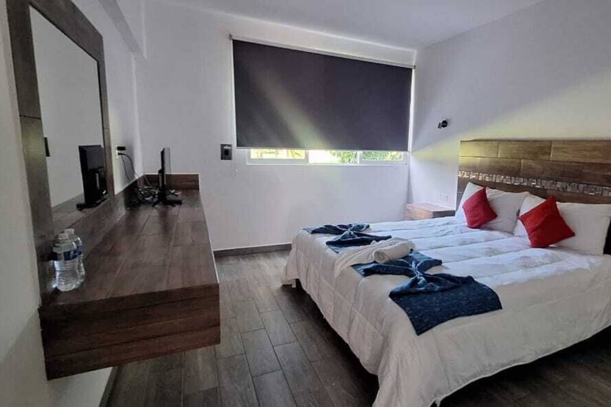 A hotel room with a bed, a desk, a chair, a mirror, a window, and a nightstand.