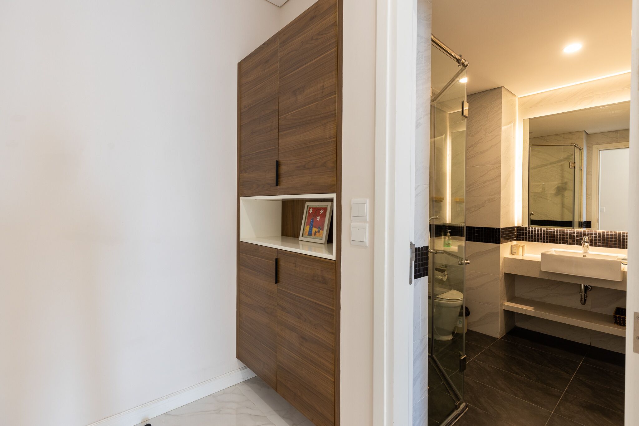 Une salle de bain moderne avec une douche à parois de verre, un lavabo blanc et une armoire en bois dotée d’une étagère.