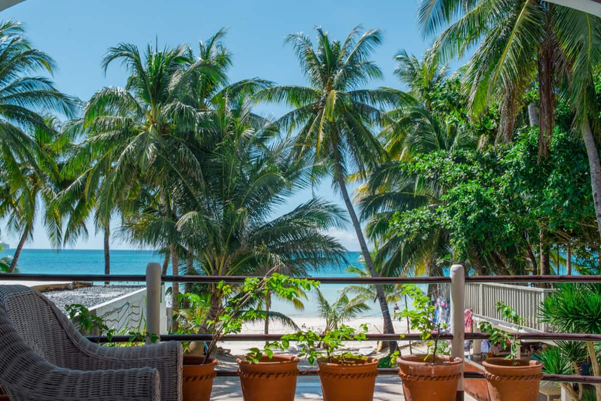 A balcony with wicker furniture and terracotta pots, overlooking a beach with palm trees and the ocean.
