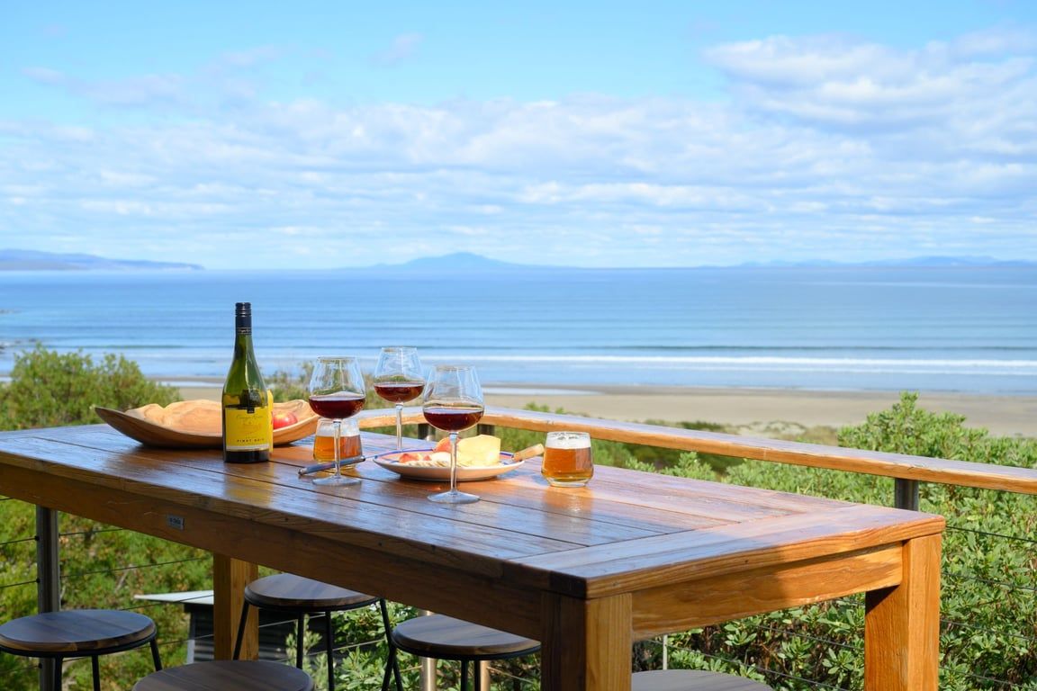 A wooden table with a bottle, glasses, and a plate of food, overlooking a beach and ocean.