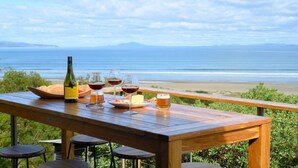 A wooden table with a bottle, glasses, and a plate of food, overlooking a beach and ocean. - Coastal Escape - Freycinet Holiday Houses (Coles Bay)