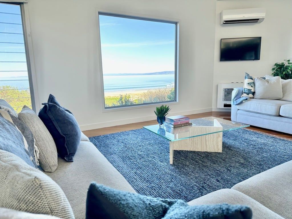 Un salon avec une grande fenêtre, un canapé blanc, une table basse en verre et un téléviseur à écran plat.
