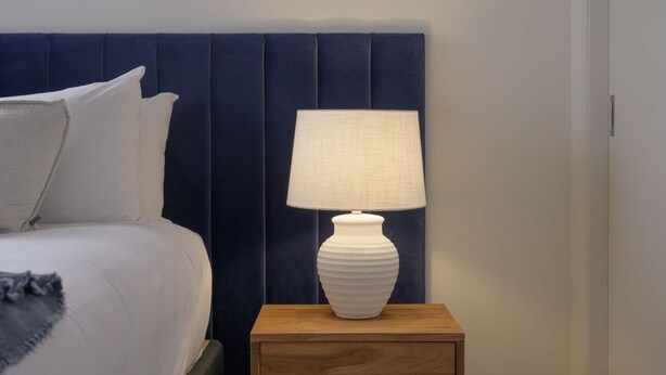 A bed with white bedding and a grey throw, a white lamp on a wooden nightstand, and a dark blue headboard. - Coastal Escape - Freycinet Holiday Houses (Coles Bay)