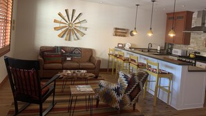 Una sala de estar con sofá, sillones, una mesa de centro y una cocina con barra y taburetes.
