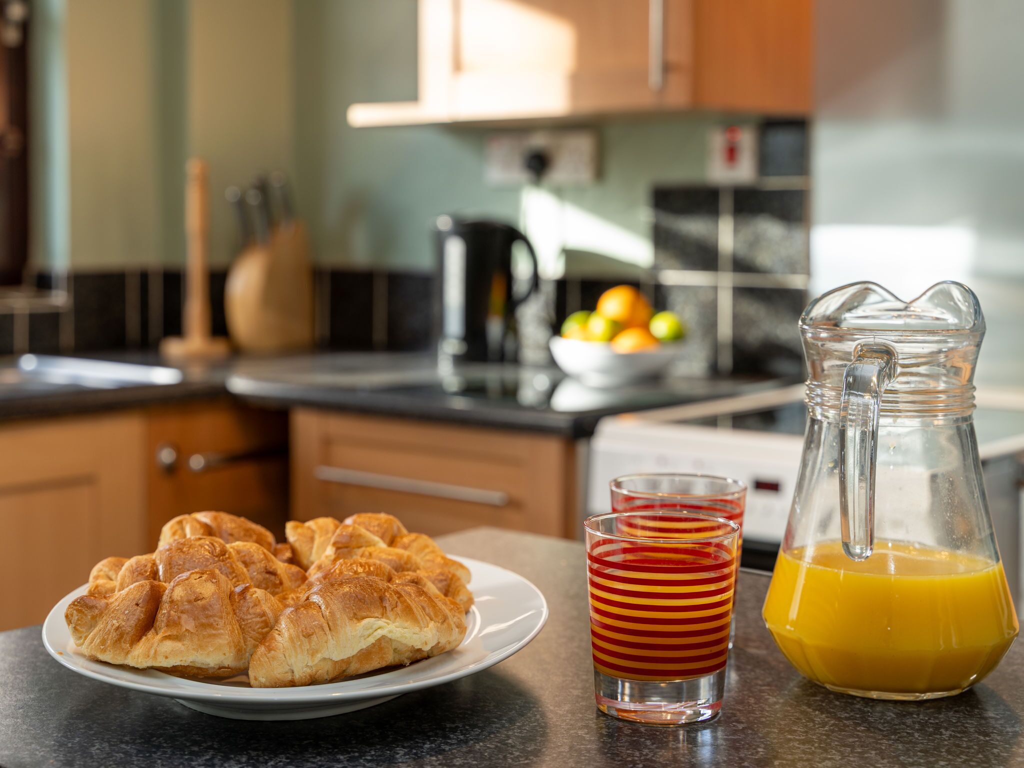 A breakfast setting with croissants on a plate, a glass of orange juice, and a pitcher of orange juice on a kitchen counter.