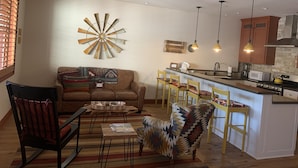 Una sala de estar con sofá, sillones, una mesa de centro y una cocina con barra y taburetes.