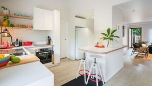 Una cocina moderna con armarios blancos, una isla central con taburetes y una zona de comedor con una silla de mimbre.