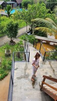 Una persona caminando sobre un sendero de concreto mojado, con una baranda metálica, rodeada de abundante vegetación y una pequeña fuente de agua.