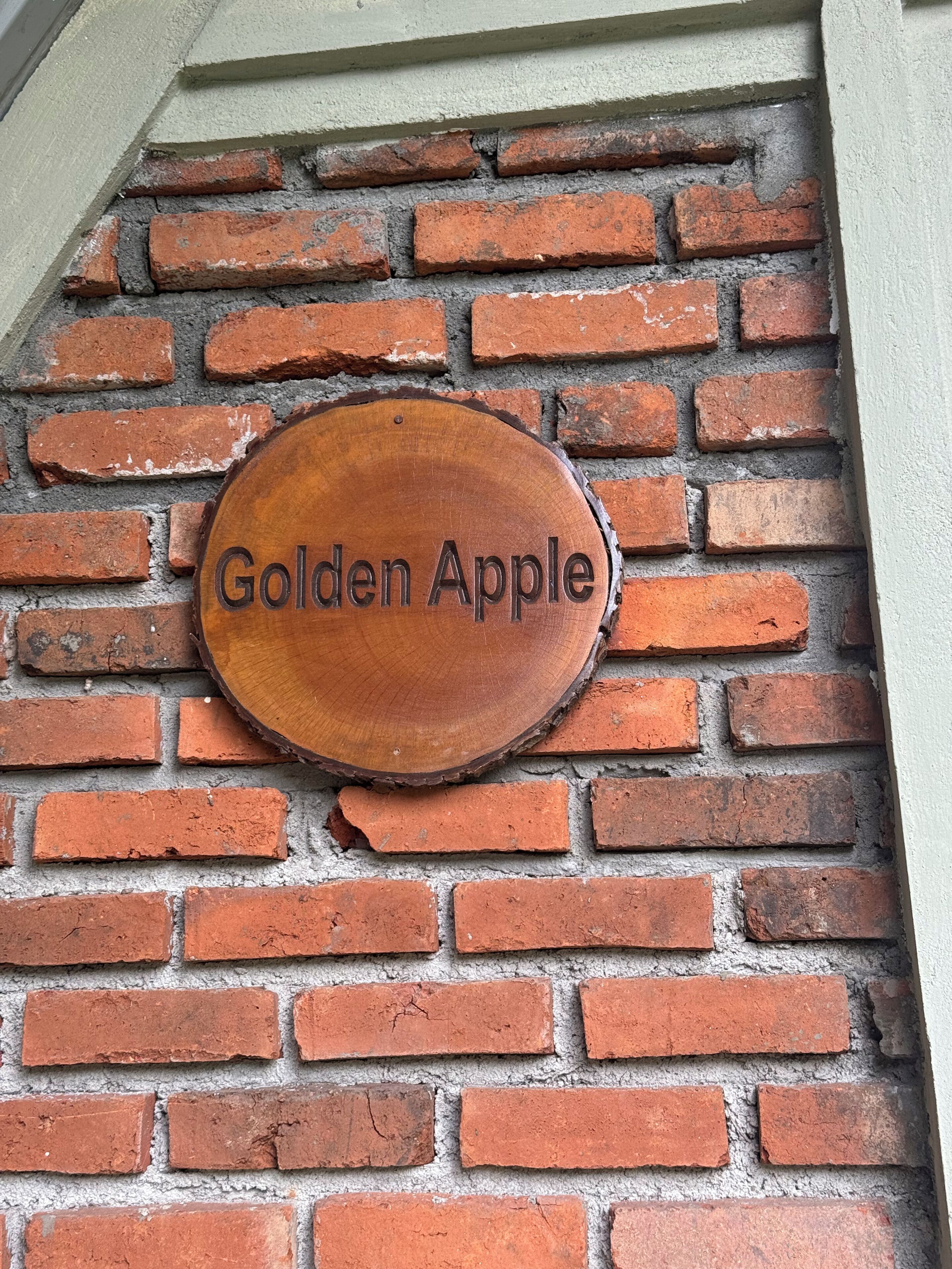 A wooden sign with "Golden Apple" engraved on it, mounted on a brick wall.