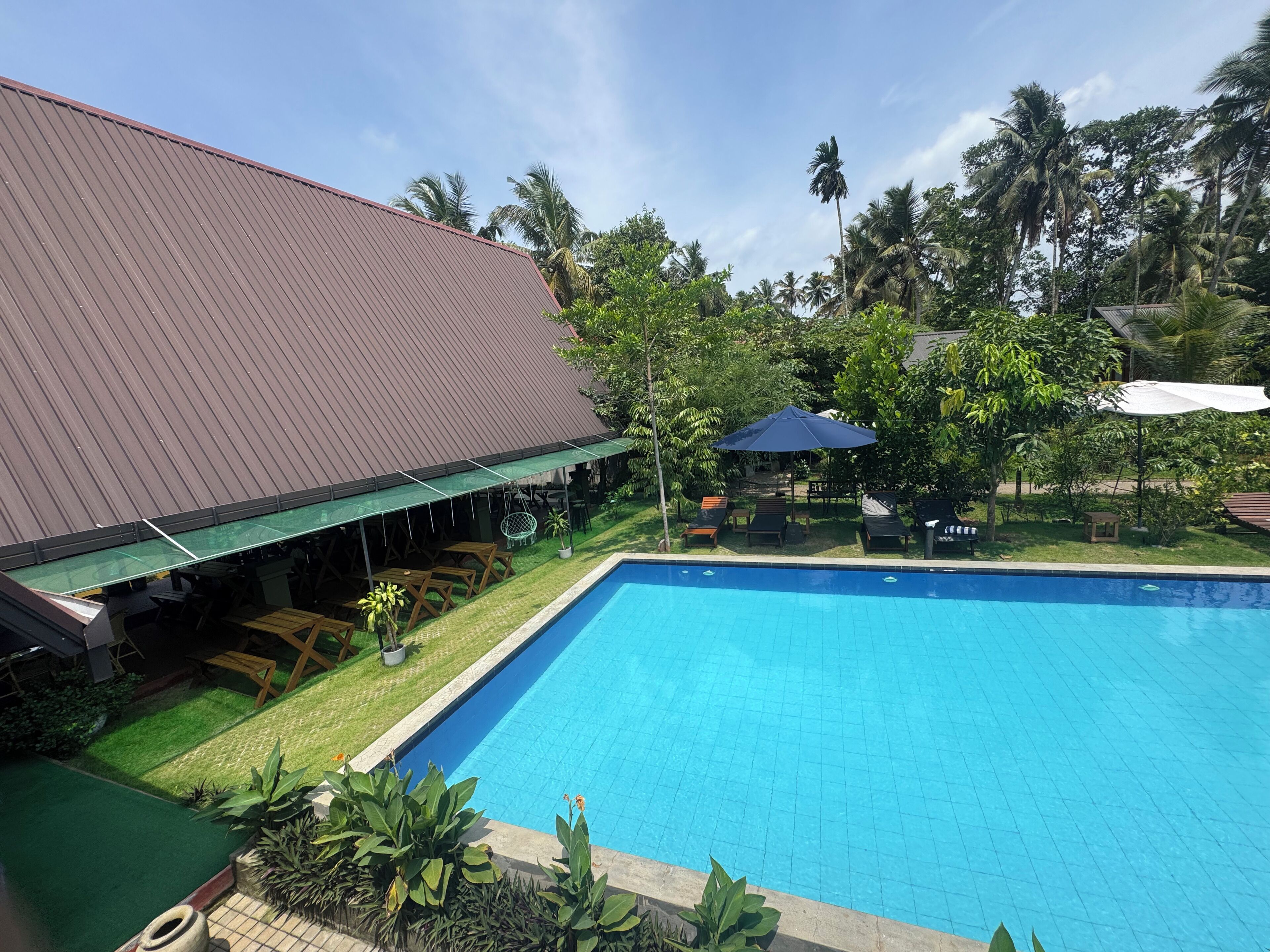 A swimming pool with a tiled deck, surrounded by greenery and a building with a red roof.