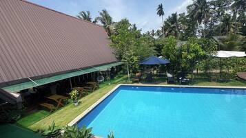 A swimming pool with a tiled deck, surrounded by greenery and a building with a red roof.