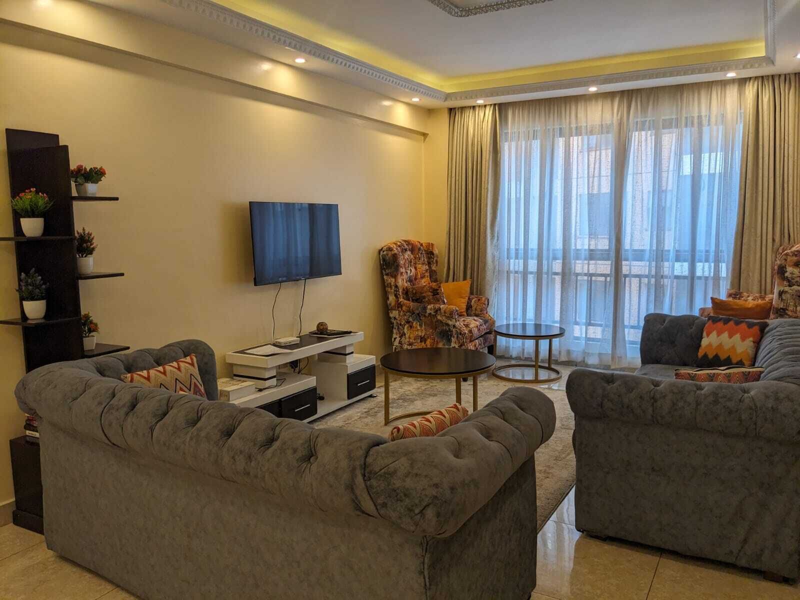 A modern living room with a large sofa, a smaller sofa, a coffee table, a TV, and a chair. There are plants on a shelf and a large window with curtains.
