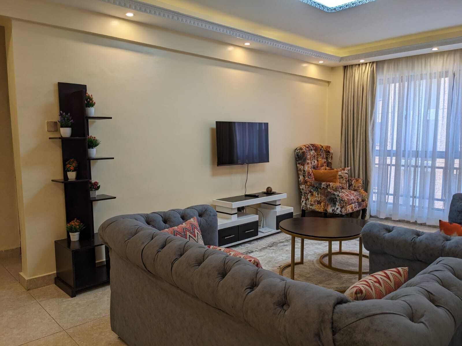 Een moderne woonkamer met een grijze bank, een fauteuil met een patroon, een flatscreen televisie, een klein bijzettafeltje en een decoratieve plank met planten.