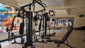 Een sportschool met diverse fitnessapparaten, een bankje en uitzicht op een speeltuin.