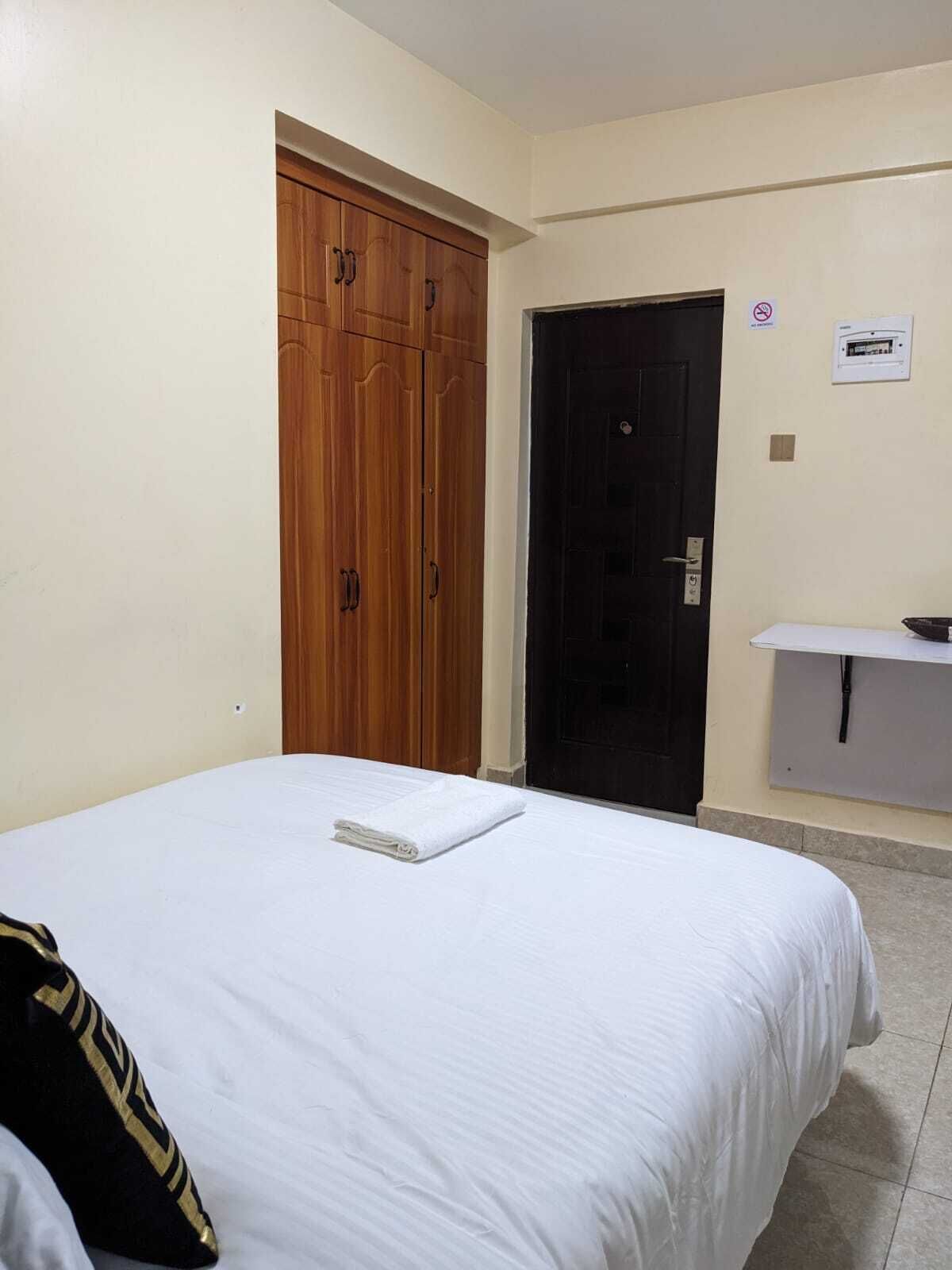 A hotel room with a bed, a wooden wardrobe, a black door, and a small white shelf.