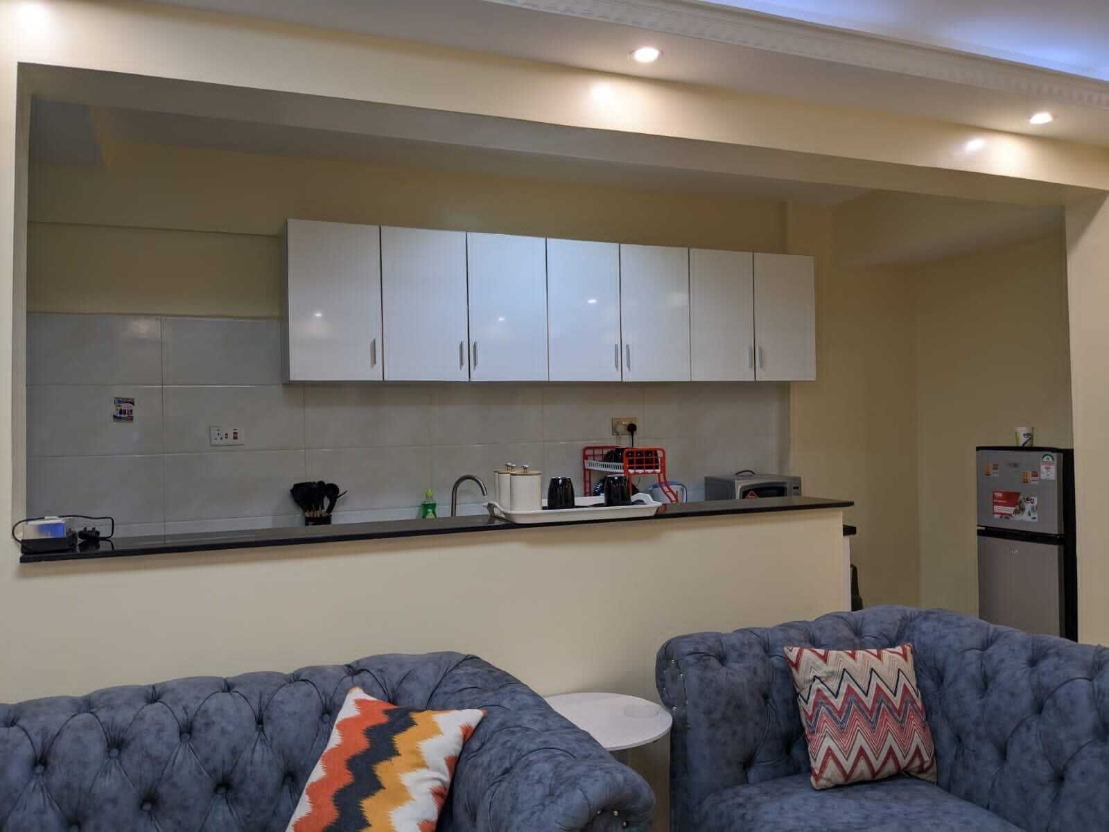 A modern kitchen with white cabinets, a black countertop, and a small round table. There are two grey upholstered sofas with patterned cushions.