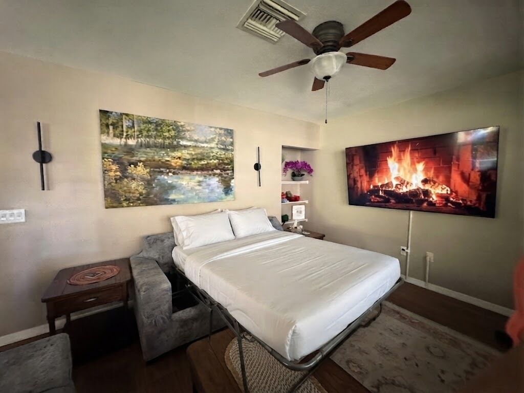 Een slaapkamer met een bed, een plafondventilator, een boslandschap op een schilderij, een schilderij van een vuur, en een nachtkastje.