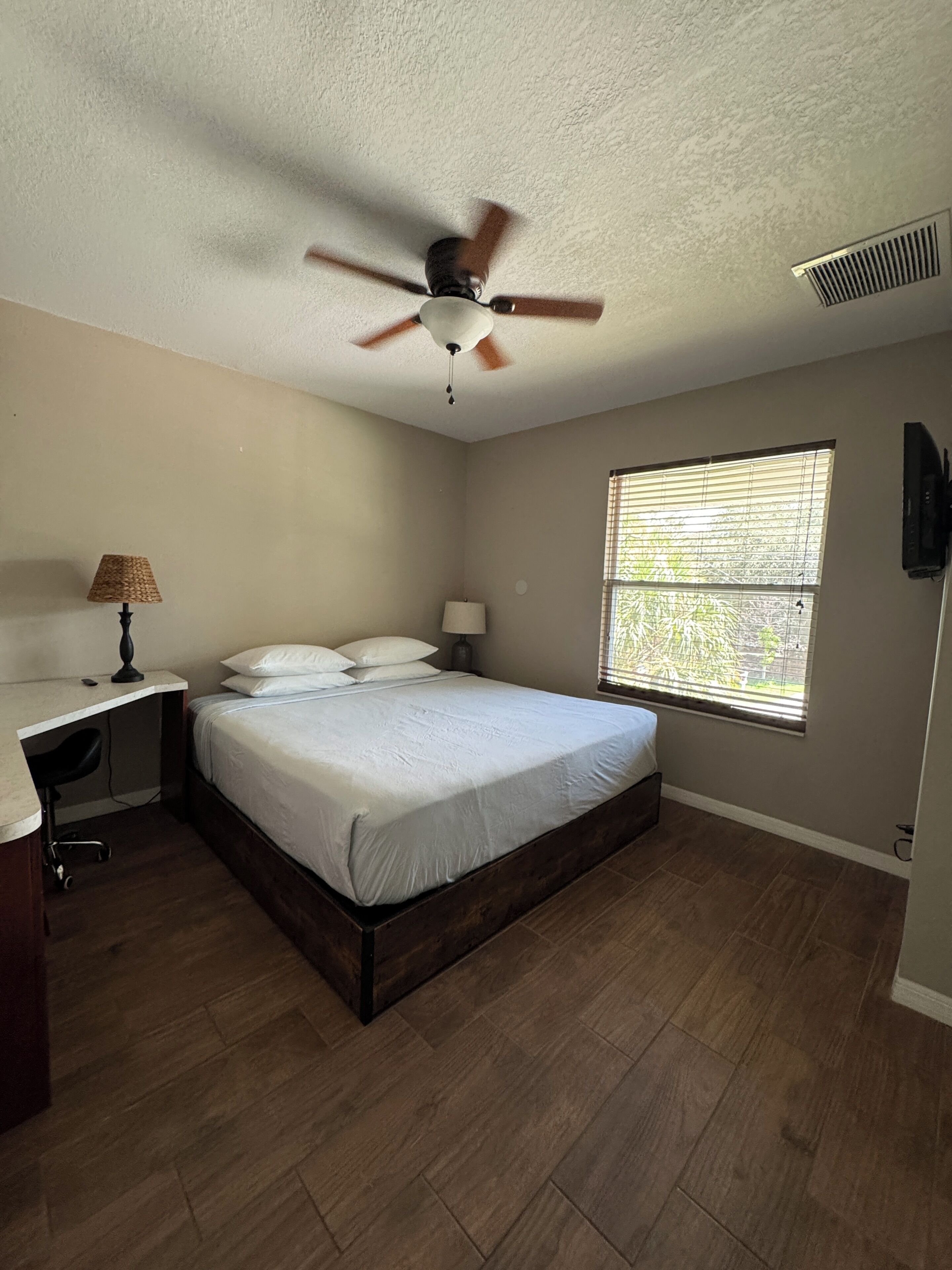 A bedroom with a bed, a desk, a chair, a ceiling fan, a window with blinds, and a TV mounted on the wall.