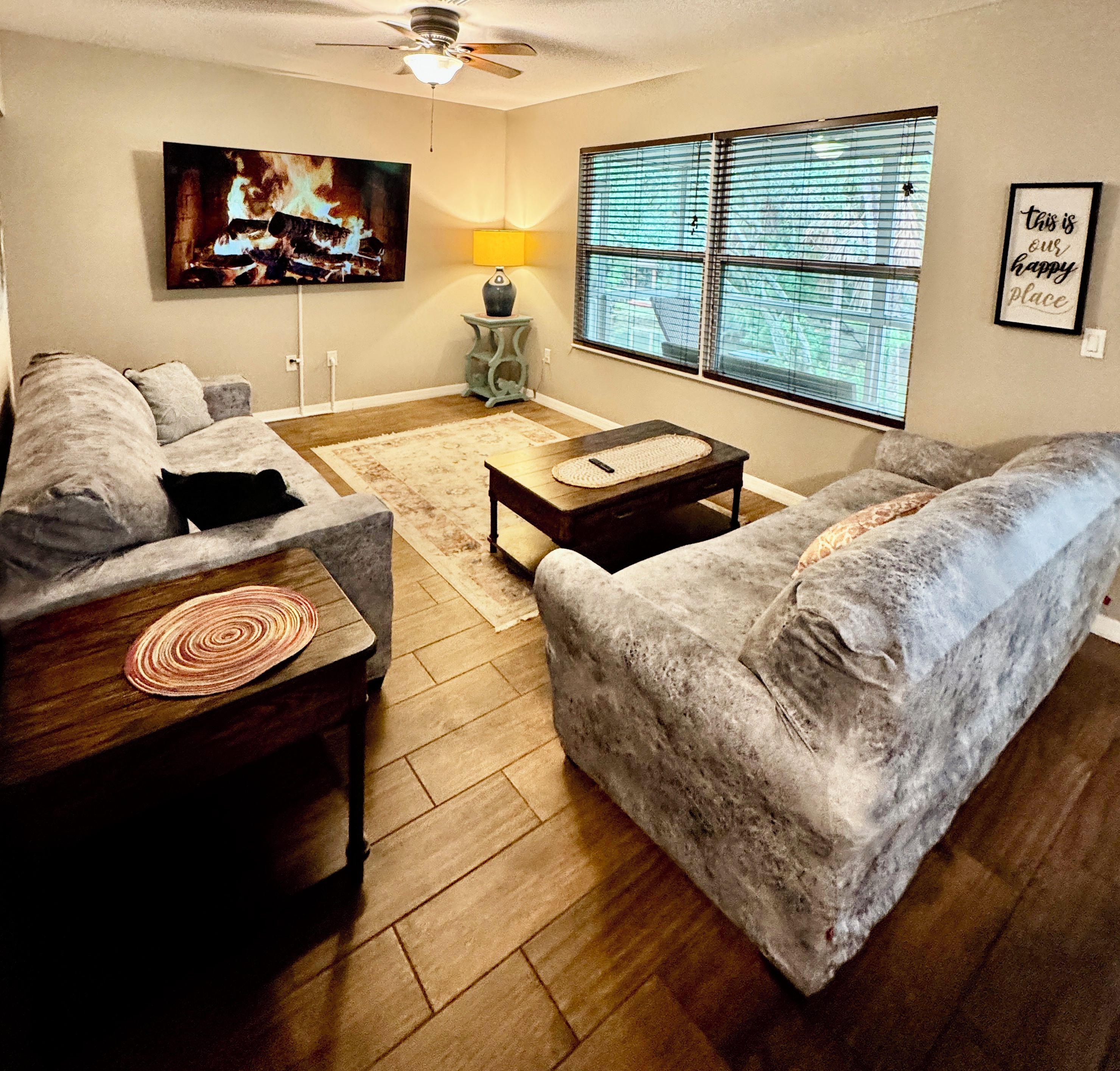A living room with a fireplace, a ceiling fan, a window with blinds, and a framed quote on the wall.