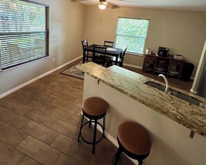 A kitchen with a granite countertop, two stools, a dining table with chairs, a window with blinds, and a ceiling fan. - Quiet getaway on a hidden Spring-run (Spring Hill)