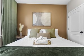 A hotel room with a large bed, green curtains, a vase with pampas grass, and a framed abstract painting on the wall.