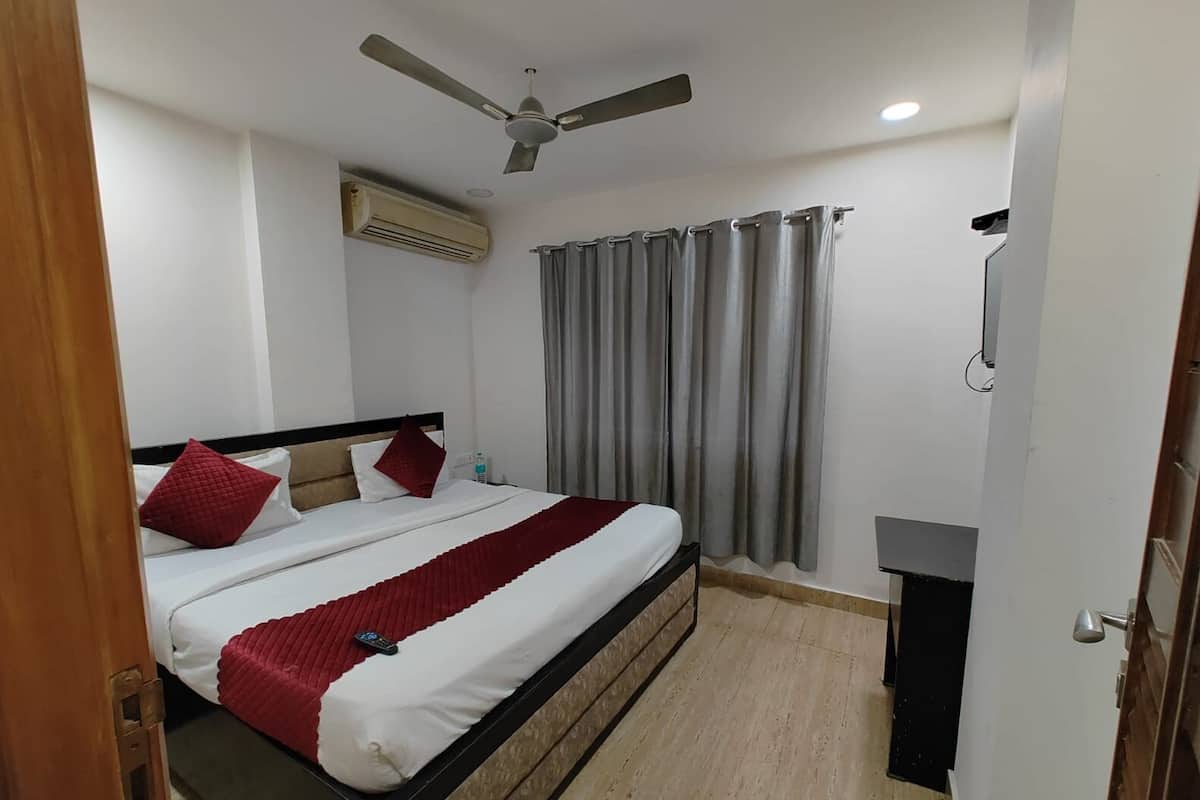 A hotel room with a bed, a ceiling fan, an air conditioning unit, a window with grey curtains, and a bedside table with a lamp.